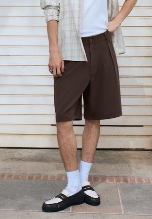 Osoba u smeđim kratkim hlačama, bijelim čarapama, crno-bijelim mokasinama, svijetlom kariranom košuljom, stoji uz bijeli zid s letvicama.