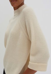 Maglione beige a costine con collo alto e maniche a tre quarti, caratterizzato da un intreccio di tessuto testurizzato e polsini arrotolati.