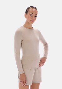 Beige pitkähihaisen paita, joka on valmistettu sileästä, kevyestä materiaalista. Siinä on pyöreä pääntie ja vartalonmyötäinen leikkaus. Yhdistetty samanlaisiin shortseihin.