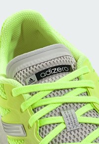 Adizero trkačka cipela s gornjim dijelom u svijetloj neon žutoj boji, sivoj mrežastoj dekoraciji i reflektirajućim detaljima na jeziku i vezicama.