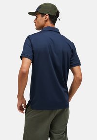 Marineblauwe polo met korte mouwen, kraag en zijsplitten. Te dragen met groene broek. Achteraanzicht toont schone lijnen en een gladde textuur.