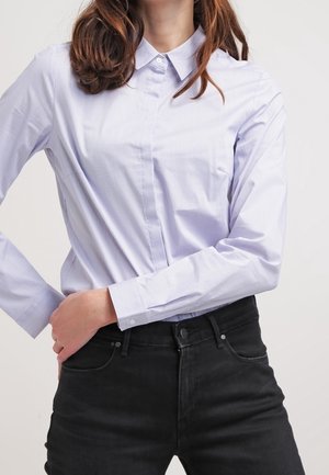 Frau trägt ein hellblaues Hemd mit Knöpfen, das in hoch taillierte schwarze Jeans gesteckt ist, mit einer Hand auf der Hüfte.