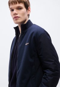 Μπλε μπουφάν με φερμουάρ, διαθέτει υφαντό σώμα με υφή και μανίκια από λείο ύφασμα, με διακριτικό λογότυπο στο στήθος.