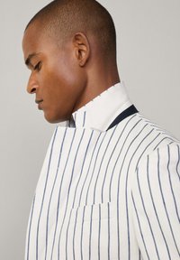 Chemise à rayures blanches et bleues, avec un col haut et une poche unique. Le tissu semble lisse avec des détails de couture visibles.