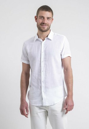 Chemise en coton blanche à manches courtes avec un devant à boutons, tissu texturé et manches roulées, associée à un pantalon de couleur claire.