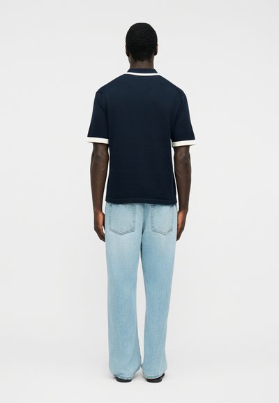 Άνδρας που στέκεται με την πλάτη σε εμάς, φορά μπλε σκούρο κοντομάνικο πουκάμισο με λευκή περίμετρο και ανοιχτόχρωμο τζιν με φαρδιά πόδια, μπροστά σε απλό φόντο.