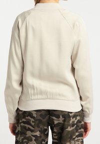 Chaqueta bomber de un beige claro, con un cuello acanalado, mangas con puños y una textura suave. Llevada sobre pantalones con patrón de camuflaje.