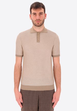 Uomo che indossa una polo beige a maniche corte con trama e patta con bottoni, pantaloni marroni, in piedi contro uno sfondo chiaro e uniforme.