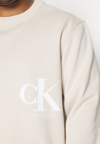 Beiger Sweatshirt mit weißem "cK"-Logo und dem Schriftzug "Calvin Klein Jeans". Weicher Stoff mit geripptem Halsausschnitt und einem klaren, minimalistischen Design.