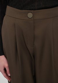 Närbild av en person som bär brun byxor med hög midja och knäppning samt en svart texturerad topp.