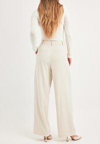 Pantalons beige à jambes larges avec une texture lisse, dotés d'une taille ceinturée, de deux poches arrière et d'une silhouette droite.