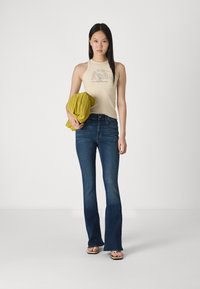 Top halter bej cu un design imprimat, blugi evazați de culoare albastru închis și un wrap texturat de culoare galbenă. Modelul stă în fața unui fundal neutru.