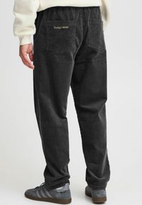 Tmavě šedé manšestrové kalhoty s vertikálním žebrováním, elastickým pasem, dvěma zadními kapsami a zlatým logem poblíž pravé kapsy.