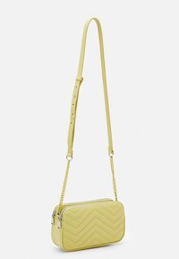 Borsa a tracolla gialla trapuntata con motivo a zigzag, chiusura con zip e tracolla a catena removibile. Realizzata in materiale sintetico con una texture liscia.