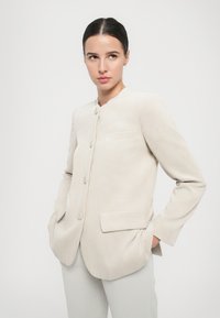 Blazer in corduroy beige chiaro con una silhouette accorciata, colletto arrotondato e due tasche frontali. Presenta bottoni grandi e una texture a coste.