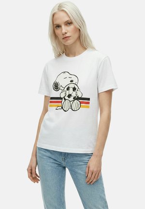 Femme blonde portant un t-shirt blanc avec Snoopy serrant un ballon de football sur des rayures noires, rouges et jaunes, associé à un jean bleu clair.