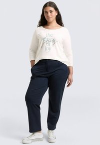 Camicia bianca a maniche lunghe con testo turchese "la stagione della gioia", abbinata a pantaloni blu navy e scarpe da ginnastica bianche, con una vestibilità comoda.