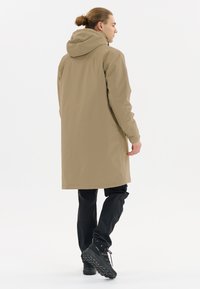 Beige waterdichte jas met een capuchon, lange ontwerp en een gladde textuur. Gecombineerd met zwarte broek en zwarte wandelschoenen.