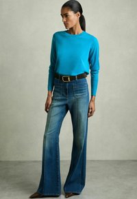 Zila garās piedurknes blūze, kas saskaņota ar augsto vidukli denim flared biksēm, ar divām priekšējām kabatām un kontrastējošu šuvju detaļām, kā arī brūnu jostu.