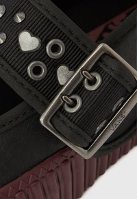 Fekete lábbeli bordázott bordó talppal, fekete pánttal, melyet ezüst szív- és fülke díszítések díszítenek, és fém csattal rögzítve.