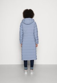 Abrigo largo acolchado de color azul claro con capucha, con diseño acolchado y puños acanalados. Llevado sobre vaqueros oscuros y zapatillas blancas.