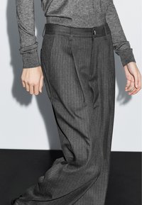 Personne portant un pantalon gris foncé à rayures fines, ample et taille haute, et un haut gris ajusté à manches longues, se tenant devant un fond clair.