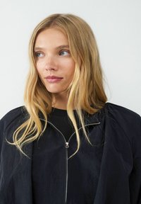 Schwarze Zip-Jacke mit weicher Textur, ausgestattet mit hohem Kragen und leicht gepufferten Schultern, über einem schwarzen Oberteil getragen.