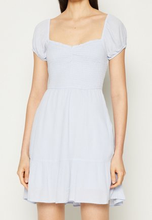 Kobieta w jasnoniebieskiej sukience z krótkim rękawem, kwadratowym dekoltem i marszczonym gorsetem, stojąca na jednolitym tle.