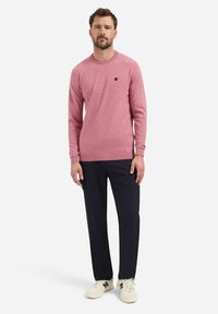 Lichtroze gebreide trui met een ronde halslijn en lange mouwen, gecombineerd met donkerblauwe broek en beige sneakers met blauwe accenten.