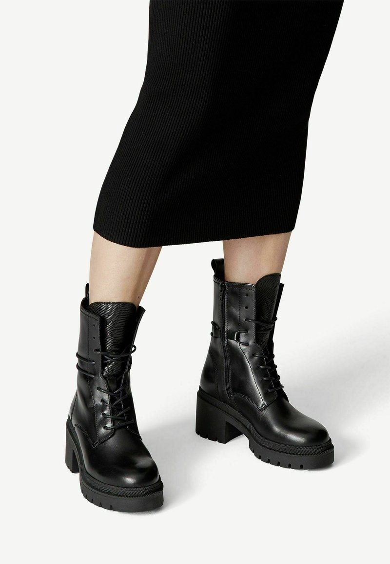 Femme portant des bottes noires en cuir à lacets mi-mollet avec talons carrés, associées à une jupe côtelée noire se terminant sous les genoux.