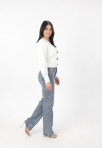 Witte geribde cardigan met knopen, gecombineerd met lichtblauwe rechte jeans en doorzichtige blokhak sandalen, tegen een witte achtergrond.