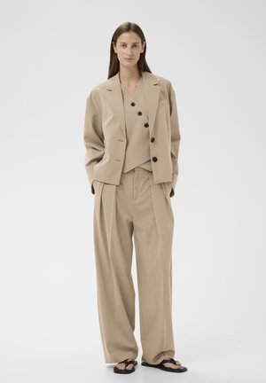 Nő bézs színű, túlméretezett, fekete gombokkal díszített öltönyben, széles szárú nadrággal, fekete szandálokkal, egyszínű fehér háttér előtt állva.