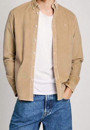 Mand iført tanfarvet skjorte med knapper over en almindelig hvid t-shirt og blå jeans, med den ene hånd i lommen, mod en neutral baggrund.
