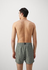 Bañadores de hombre en color verde oliva con un patrón floral, cinturilla elástica y un bolsillo trasero con parche. Textura de tela suave.