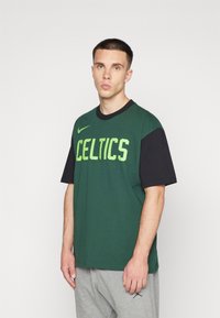 La camiseta Nike en verde y negro presenta "CELTICS" en letras verde brillante, con un corte relajado y mangas cortas, combinada con pantalones grises.