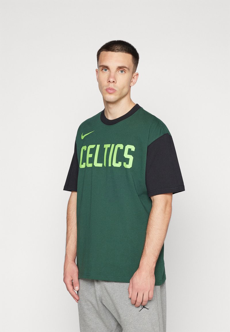 La camiseta Nike en verde y negro presenta "CELTICS" en letras verde brillante, con un corte relajado y mangas cortas, combinada con pantalones grises.