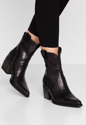 Femme portant des bottines noires texturées à bouts pointus et talons blocs, associées à un pantalon noir ajusté.