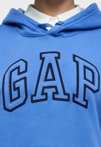 Άτομο που φοράει μπλε φούτερ με κουκούλα με μεγάλα ραμμένα γράμματα "GAP" πάνω από λευκό πουκάμισο με γιακά, κοντινή λήψη στο πάνω μέρος του στήθους και τον λαιμό.