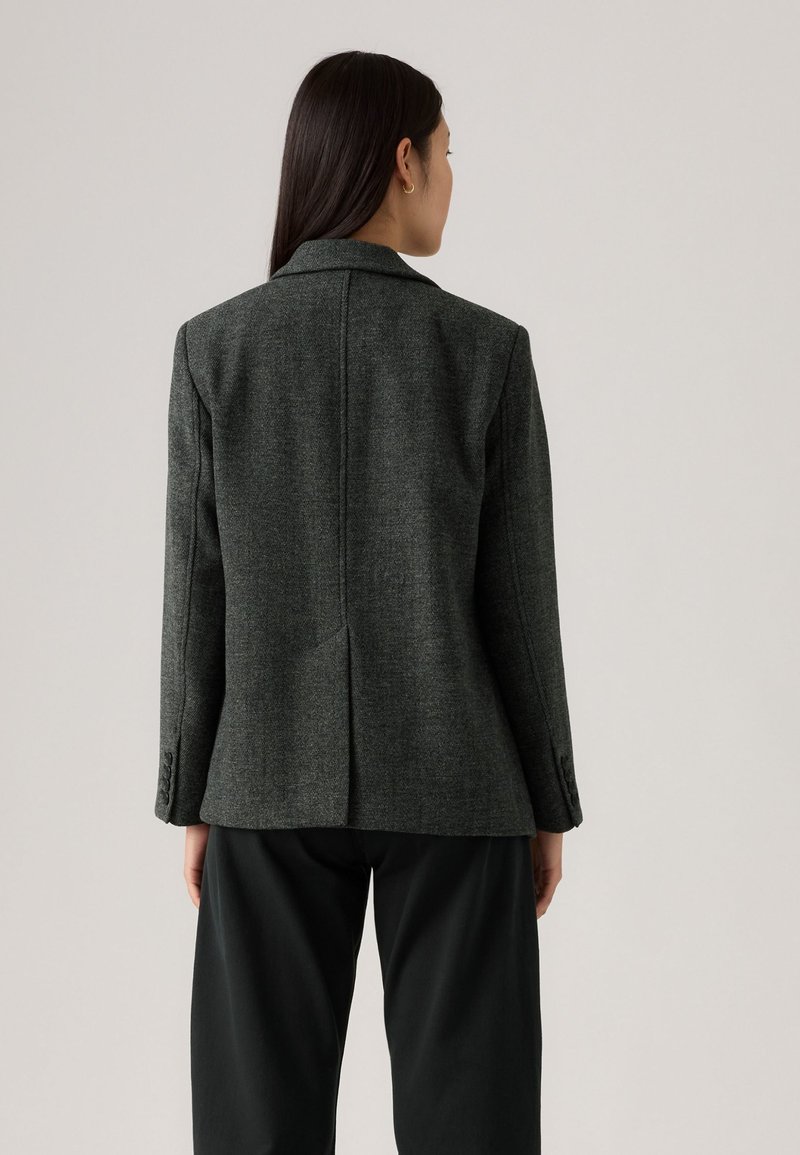 Mörkgrå skräddarsydd blazer med en strukturerad silhuett, snygg krage, en slits bak och tre-knappsribbor. Bärs över svarta byxor.