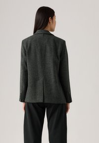Blazer verde scuro su misura con tessuto strutturato, rever a lancia, spacco singolo sul retro e polsini con bottoni, abbinato a pantaloni neri.