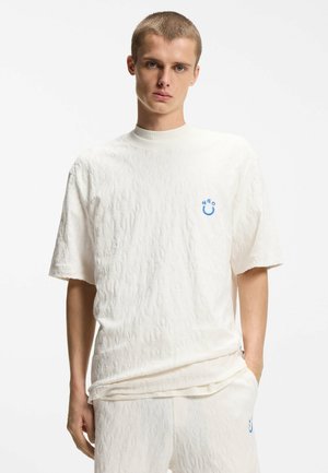 Giovane con capelli corti che indossa una camicia bianca a maniche corte con trama e pantaloncini abbinati, entrambi con un piccolo logo blu a forma di faccina sorridente sul petto.