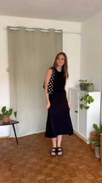 Svart ärmlös klänning med rak fåll, kombinerad med en vit väst med prickigt mönster, svarta sandaler och minimala accessoarer.
