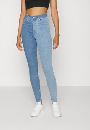 Femme portant un jean skinny taille haute avec une jambe bleu foncé et l'autre bleu clair, assorti à des baskets blanches et un haut court noir.