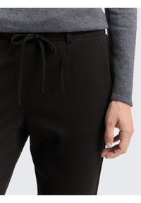 Pantaloni neri con una texture liscia, dotati di vita regolabile con cordino e tasche laterali, abbinati a una maglietta a maniche lunghe grigia chiara.