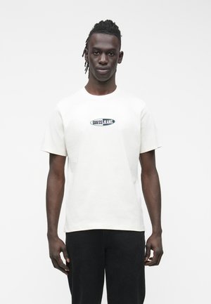 T-shirt blanc en coton avec col rond et manches courtes, arborant un logo "GUESS JEANS" vert brodé sur la poitrine.