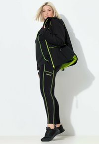 Chaqueta deportiva negra con acentos verdes, combinada con leggings negros con franjas laterales verdes. Modelo lleva zapatillas negras.