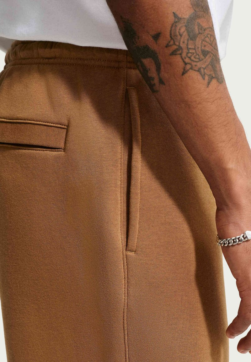 Pantalon de survêtement marron avec une texture lisse, doté d'une taille élastique, d'un cordon de serrage et d'un détail de poche arrière.