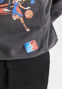 Sudadera gris con un gráfico colorido de un jugador de baloncesto. Incluye una etiqueta de logo multicolor en el dobladillo. Pantalones negros texturizados visibles abajo.