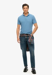 Polo shirt azzurro chiaro, jeans blu scuro e mocassini neri; camicia a scacchi legata in vita; indossa un orologio al polso sinistro.