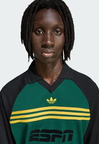 Zelena i crna polo majica sa žutim prugama, s logotipom Adidas i natpisom 'ESPN'. Tkanina izgleda teksturirano i prozračno.
